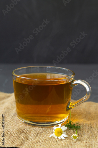 hot drinks. mug of chamomile tea on sackcloth. glass cup of iced Herbal chamomile tea on a black table with copy space. cup of tea with fresh flowers and green leaves on grey background. vertical