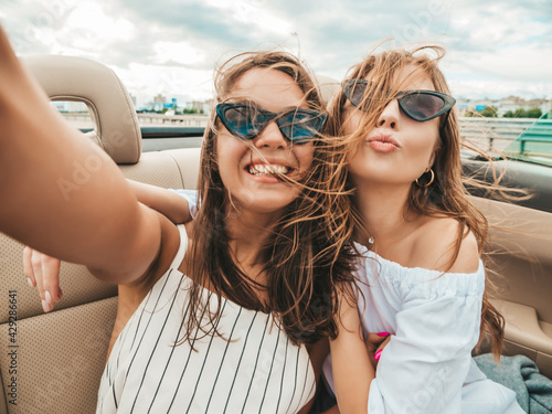 Portrait of two young beautiful and smiling hipster female in convertible car. Sexy carefree women driving cabriolet. Positive models riding and having fun in sunglasses outdoors.Enjoying summer days