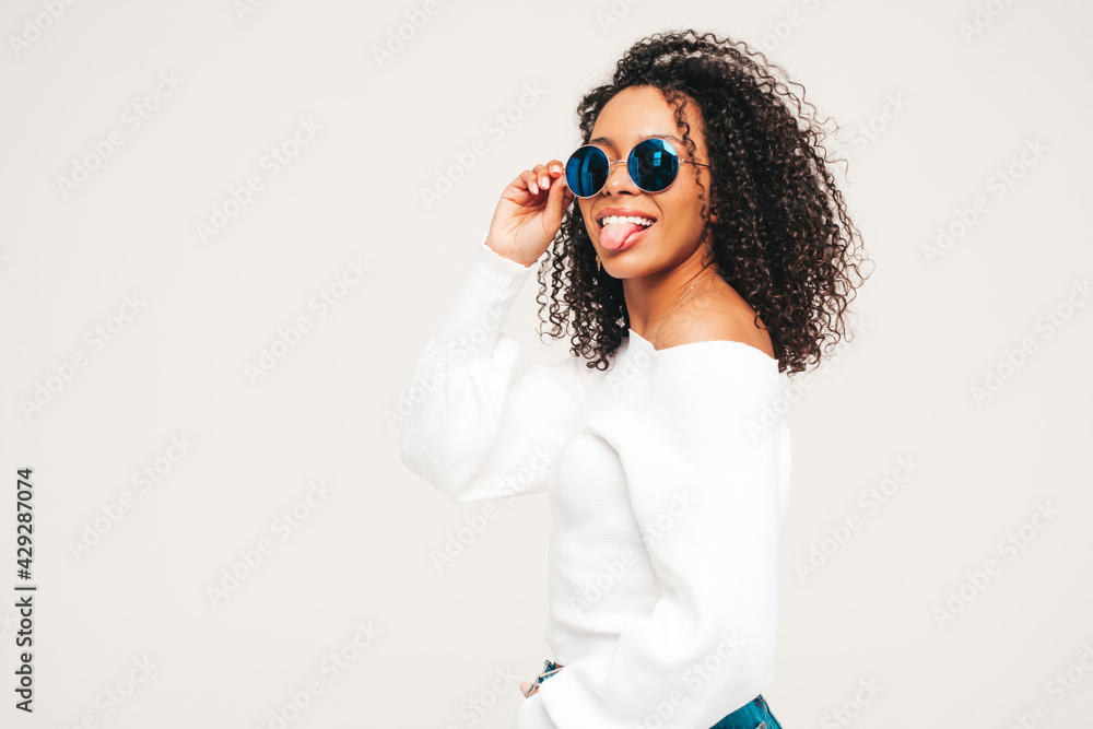 Beautiful black woman with afro curls hairstyle.Smiling model in sweater and trendy jeans clothes. Sexy carefree female posing on white background in studio. Tanned and cheerful. In sunglasses Beautiful black woman with afro curls hairstyle.Smiling model in sweater and trendy jeans clothes. Sexy carefree female posing on white background in studio. Tanned and cheerful. In sunglasses