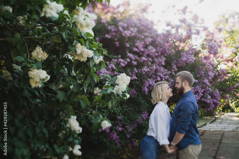 Romantic and happy caucasian couple in casual clothes hugging on the background of beautiful blooming lilac. Love, relationships, romance, happiness concept. Man and woman walking outdoors together.