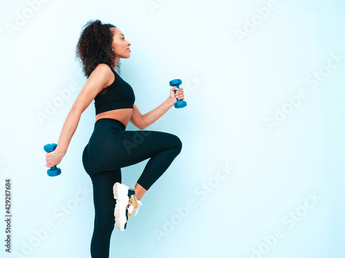 Fitness smiling black woman in sports clothing with afro curls hairstyle.She running and jogging . Young beautiful model with perfect tanned body.Female holding dumbbells in studio near blue wall