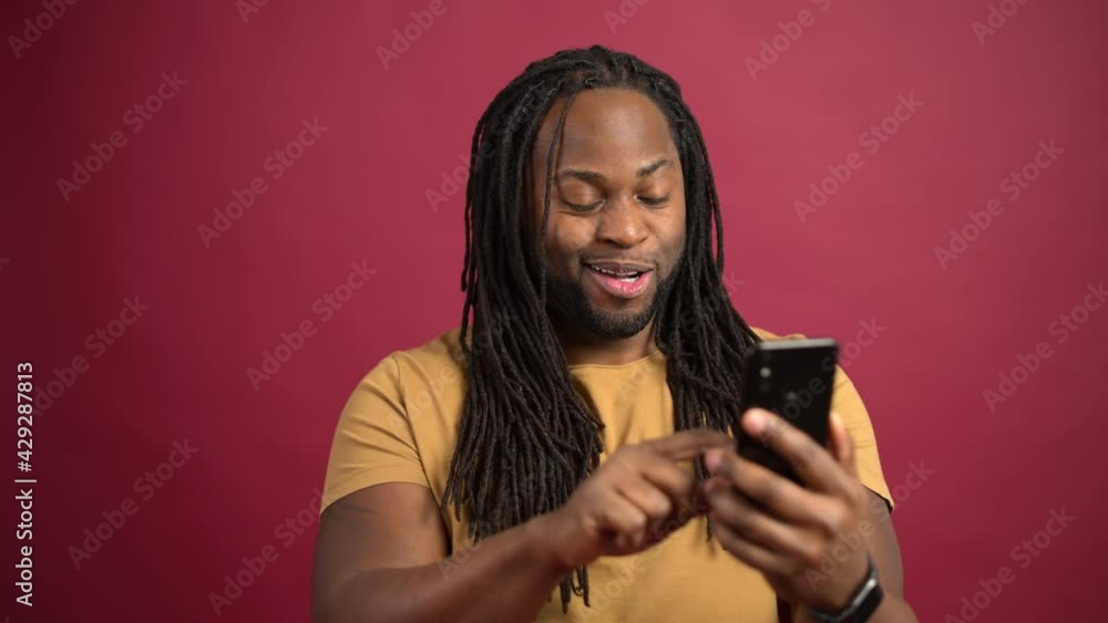 Cheerful African-American man with dreadlocks enjoys spending time online in smartphone, happy black guy scrolling news feed in social media, chatting with friends, using mobile phone for web surfing Cheerful African-American man with dreadlocks enjoys spending time online in smartphone, happy black guy scrolling news feed in social media, chatting with friends, using mobile phone for web surfing