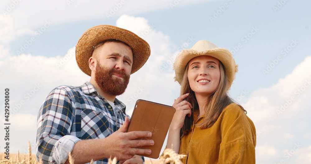 Portrait of joyful Caucasian handsome man and beautiful woman standing outdoors on sky background. Male in hat tapping and browsing on tablet in field. Girl looking far away. Couple concept