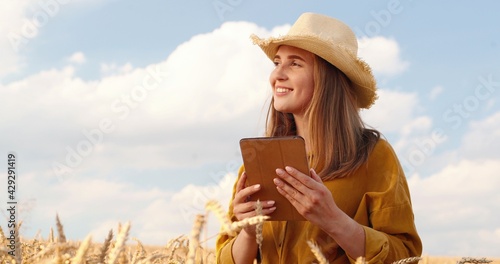 Portrait of happy pretty Caucasian young woman in hat smiling and typing on tablet while standing in wheat field and looking away. Beautiful joyful girl texting on device in village. Non-urban concept photo