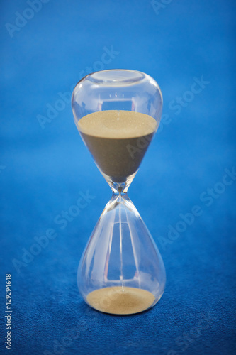 Brown sand trickles through an hourglass that stands on a blue background. You neither stop the time, nor freeze it. It goes on and on. Daylight. Indoor. Normal perspective. 
