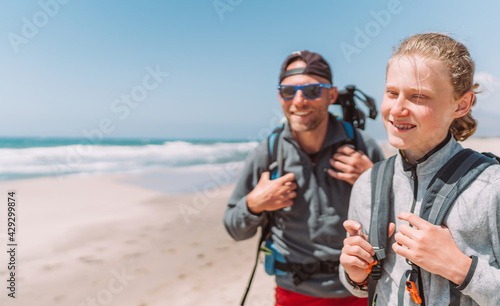 Father with teenager son with backpacks walking by the sandy seaside beach. They smiling and looking at the camera. Active happy family people vacation time concept image.