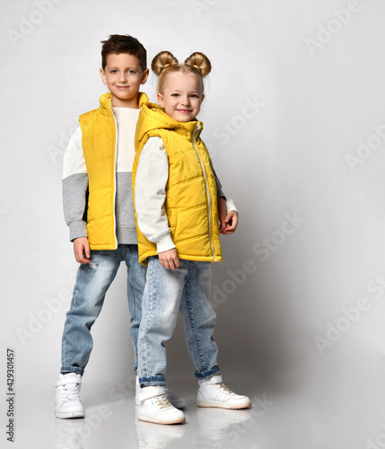 teenage boy and girl posing against a light background, children are dressed in yellow puffy sleeveless zipper vests and jeans. urban teen fashion