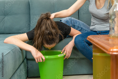Dark-haired young woman is lying on the sofa, she is ill from alcohol intoxication, she is going to vomit over a bucket, a friend came to her aid, holds her by the hair, empty bottles are poison