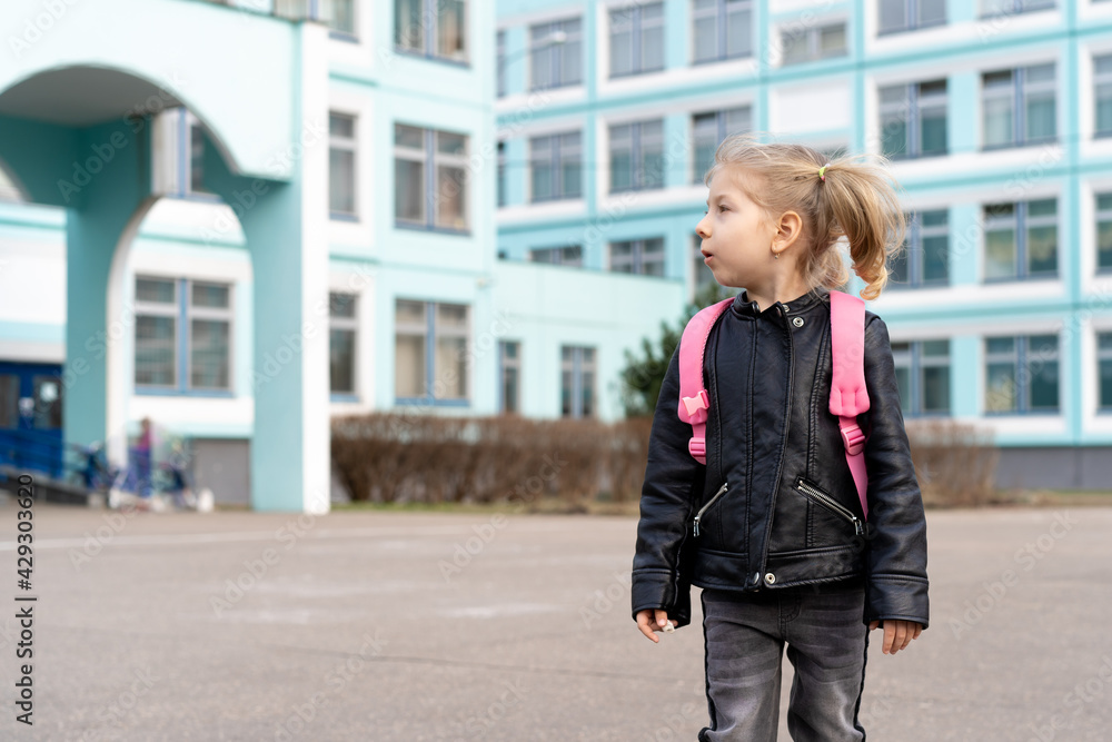Happy little girl first-grader stands near school with a pink backpack and smiles Happy little girl first-grader stands near school with a pink backpack and smiles