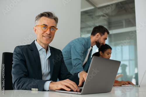 Confident mature business man wearing stylish eyeglasses using laptop computer looking at camera. Group of business people working together in modern office  © ARUTA Images