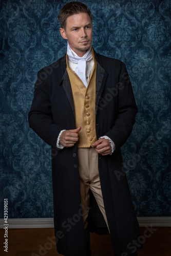 A handsome Regency gentleman wearing a gold waistcoat, breeches, and a black long jacket and standing in a room with blue wallpaper and a wooden floor photo