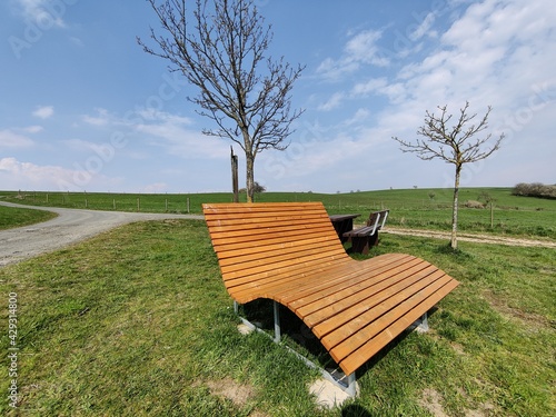 Luxuriöse Sonnenliege im Frühling oder Sommer bei Sonne, blauem Himmel und weißen Wolken an einer Wegkreuzung mit zwei Bäumen