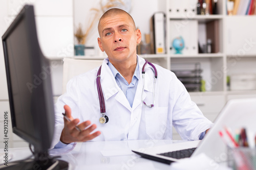 Positive man doctor sitting at workplace with computer in her office