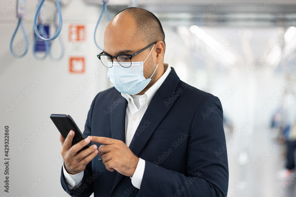 Young asian businessman wearing protection mask using smartphone standing in skytrain. Businessman in urban city travel by sky train. Young asian businessman wearing protection mask using smartphone standing in skytrain. Businessman in urban city travel by sky train.