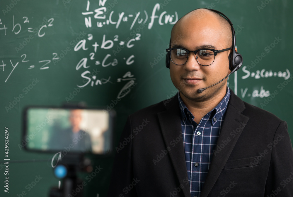 Indian young teacher man wearing headset teaching online video conference live stream by smartphone. Asian teacher teaching mathematics class webinar online for students learning. Indian young teacher man wearing headset teaching online video conference live stream by smartphone. Asian teacher teaching mathematics class webinar online for students learning.