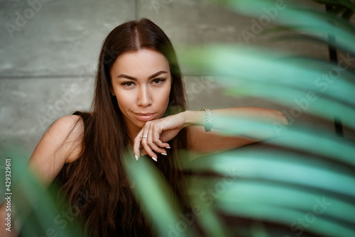 Beautiful young woman closeup portrait through green leaves of a flower on a gray wall background