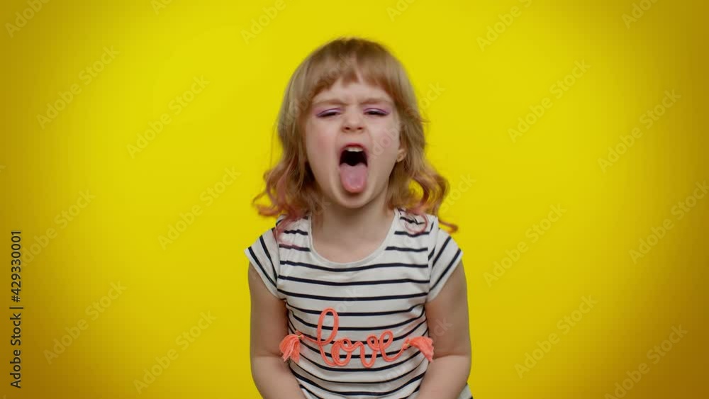 Portrait of cheerful funny blonde child girl 5-6 years old showing tongue making faces at camera, fooling around, joking, aping with silly face, teasing. Yellow studio background. Teen kid children