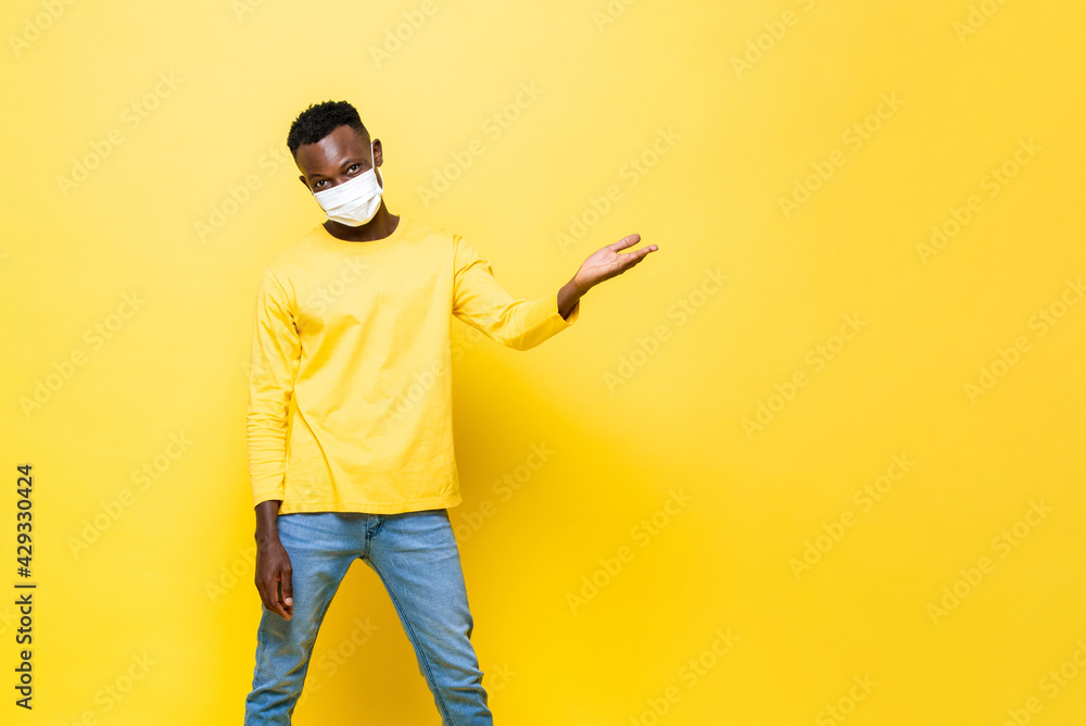 Portrait of young African man wearing face mask protecting from Corovavirus with hand opening to empty space aside in isolated yellow studio background Portrait of young African man wearing face mask protecting from Corovavirus with hand opening to empty space aside in isolated yellow studio background