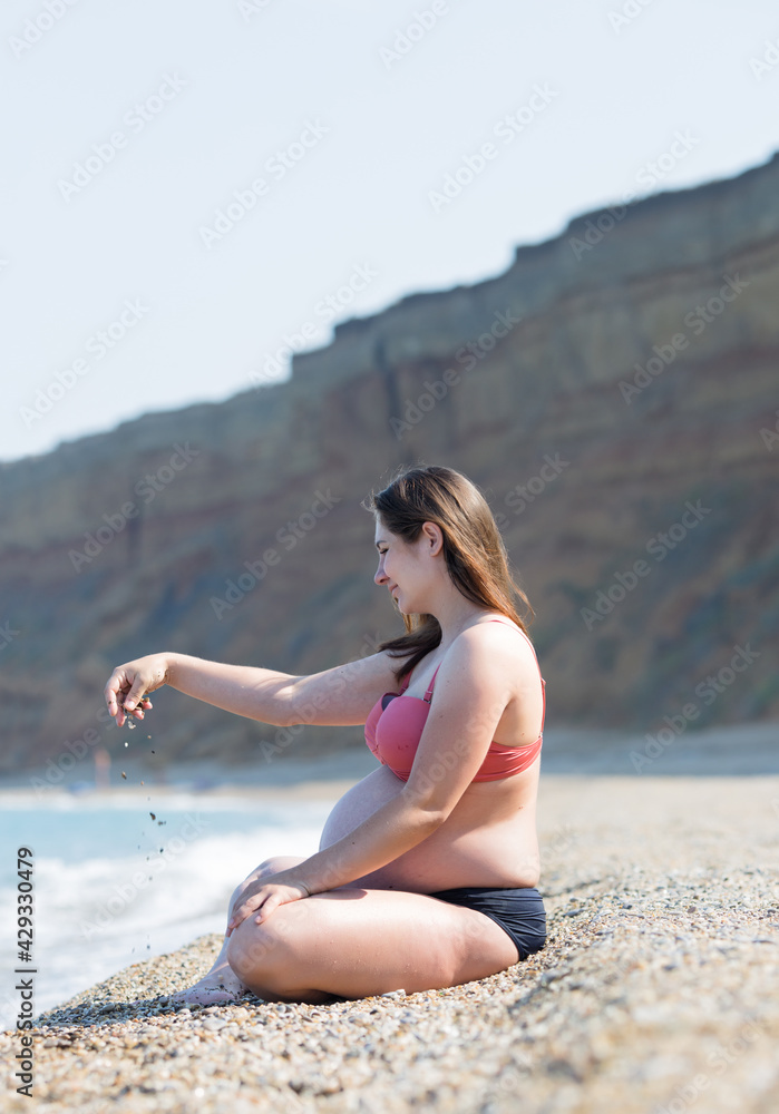 Pregnant woman in bikini resting on pebble beach in early time