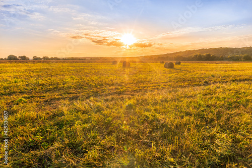 Scenic view at picturesque burning sunset in a green shiny field with hay stacks  bright cloudy sky   trees and golden sun rays  summer valley landscape