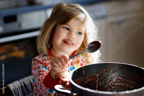 Cute toddler girl eating chocolate dough rests with spoon and fingers from pot. Happy child licking sweet dough for muffins or cake, helping in home kitchen, indoors.