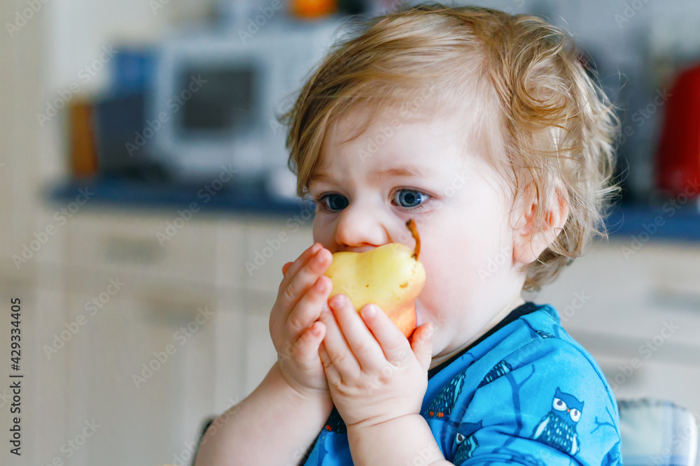 Cute adorable toddler girl eating fresh pear. Hungry happy baby child of one year holding fruit. Girl in domestic kitchen, having healthy meal snack. Smiling blond toddler at home or nursery Cute adorable toddler girl eating fresh pear. Hungry happy baby child of one year holding fruit. Girl in domestic kitchen, having healthy meal snack. Smiling blond toddler at home or nursery