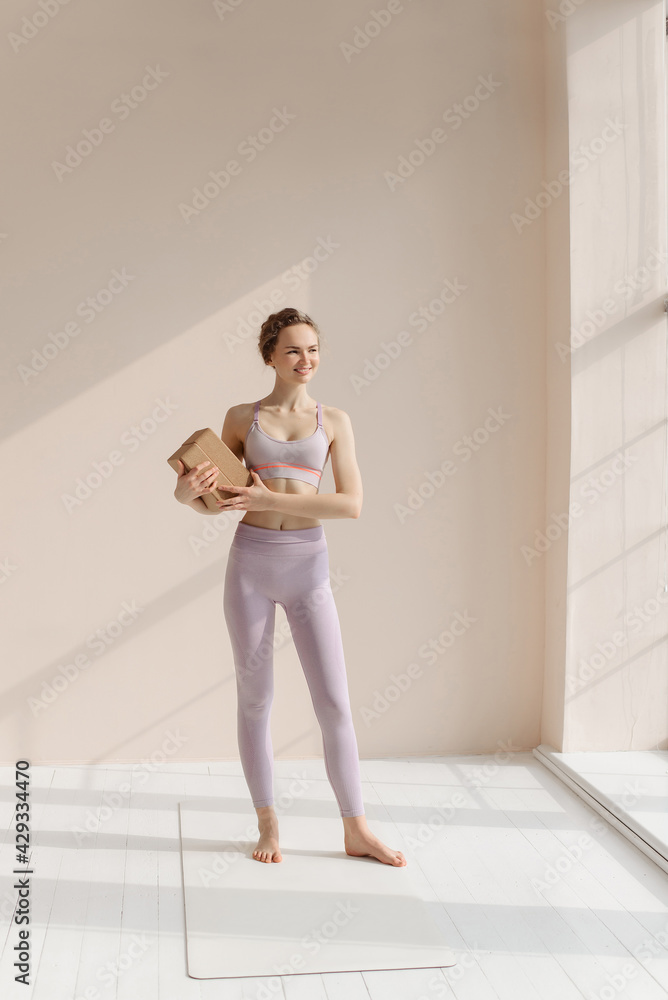 The girl does yoga. Pilates block yoga cork brick. A young woman in a sports suit on a beige one-ton background smiles.