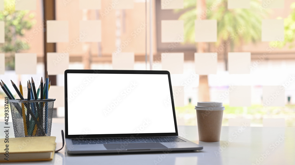 Obraz premium Open computer laptop with blank screen, notebook, coffee cup and pencil holder on white table near window.