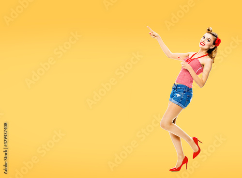 Full body portrait - amazed happy woman pointing at something. Excited girl in pin up style, showing copy space for text or imaginary. Retro fashion and vintage. Orange yellow color background.