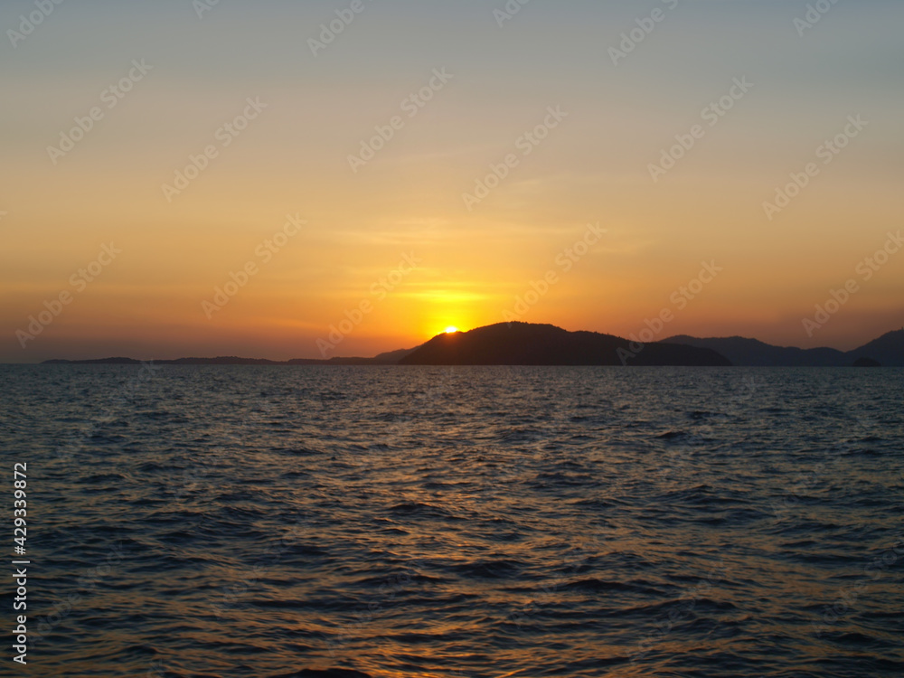 Sunset over the sea. Island on the horizon. Yellow orange glow on a clear sky. Ripple on the surface of the water. Waves, ocean, expanse of water. View from the sea, land in the distance. Andaman sea