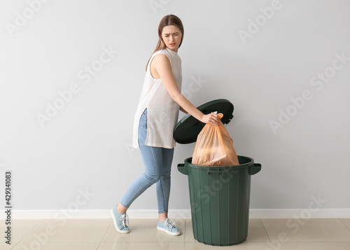 Young woman putting garbage in trash bin on light background