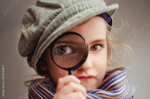 A girl in the guise of a detective looks through a magnifying glass. She has an enlarged eye. Joke and humor concept