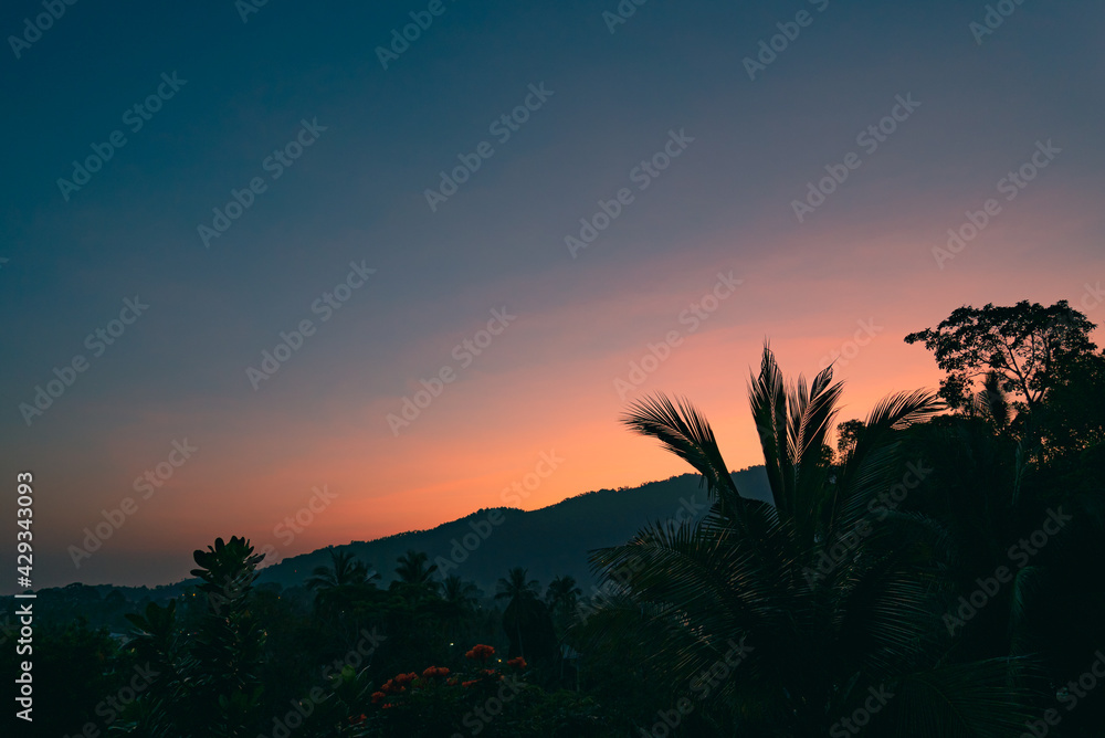 Colorful sunset sky over silhouettes of a trees and mountain on a tropical island. View from balcony.
