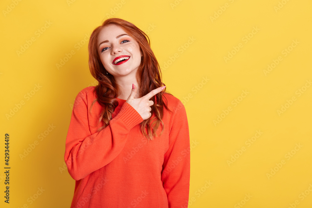 Portrait of young woman with red hair pointing index finger aside, showing to copy space for promotion, wearing casual sweater, looking at camera with happy facial expression, isolated. Portrait of young woman with red hair pointing index finger aside, showing to copy space for promotion, wearing casual sweater, looking at camera with happy facial expression, isolated.