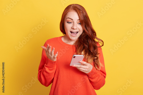 Excited red haired woman looking at screen of smart phone with surprised facial expression, girl winning lottery, receives great news, posing isolated over yellow background. © sementsova321