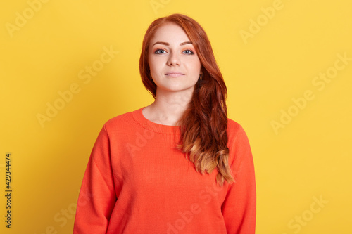 Front view of serious young attractive woman with red hair wearing casual orange sweater, looking directly at camera with confident expression, isolated over yellow background.