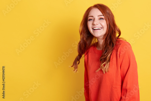 Canvastavla Portrait of young happy adorable woman with red hair dresses sweater posing isolated over yellow background and laughing happily, copy space for advertisement