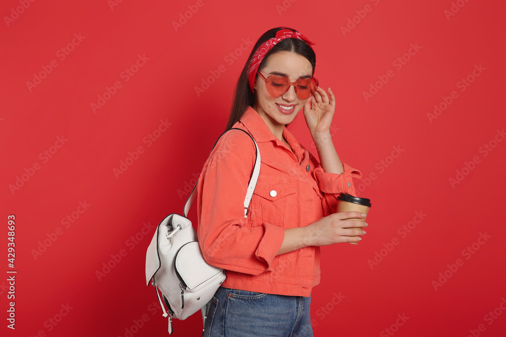 Beautiful young woman with stylish leather backpack and cup of coffee on red background Beautiful young woman with stylish leather backpack and cup of coffee on red background