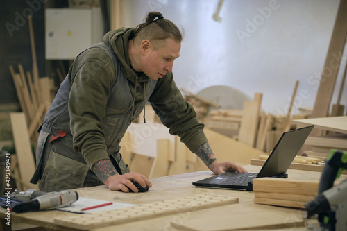 Concentrated adult craftsman checking information on laptop while working in carpentry workshop. profession, carpentry, technology and people concept photo