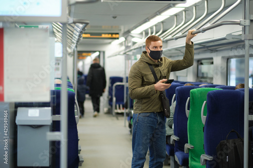 Handsome guy in respirator use a smartphone and keep social distancing to crowd while commuting in the metro or train. Infection and air pollution prevention concept © yavdat