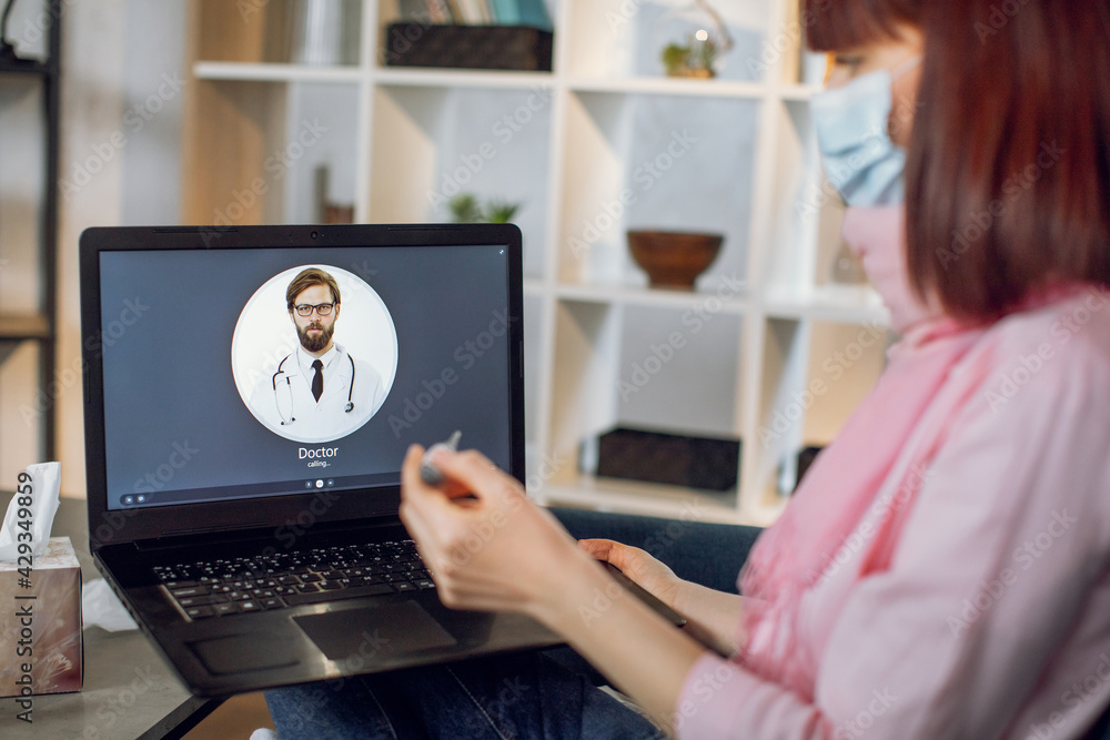 Fototapeta premium Stay at home concept. Covid prevention. Ill woman sitting on sofa at home, feeling bad and having fever, holding thermometer and calling to a doctor using laptop. Online consultation with doctor.