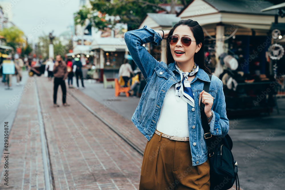 asian tourist wearing sunglasses and pushing hair back is strolling on street with metal tracks in road. blurred background happy stylish lady enjoying the beautiful scenery of the simple town