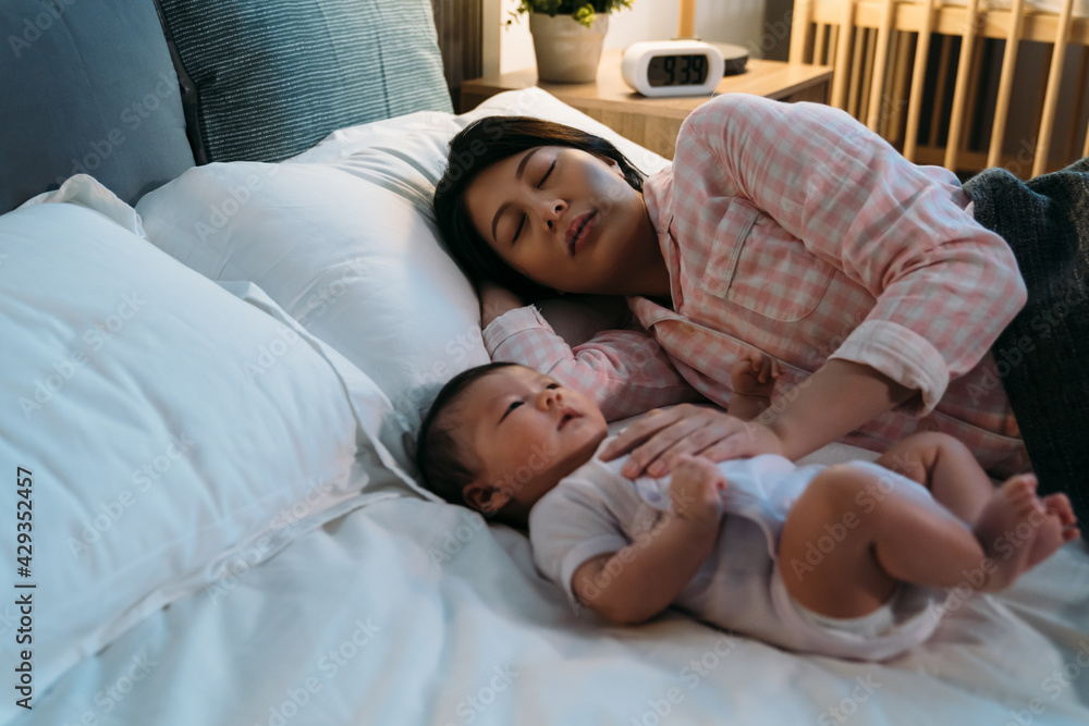 tired asian new mom wearing pajamas is falling asleep on the bed while comforting her waking baby at night during bedtime in the bedroom. tired asian new mom wearing pajamas is falling asleep on the bed while comforting her waking baby at night during bedtime in the bedroom.