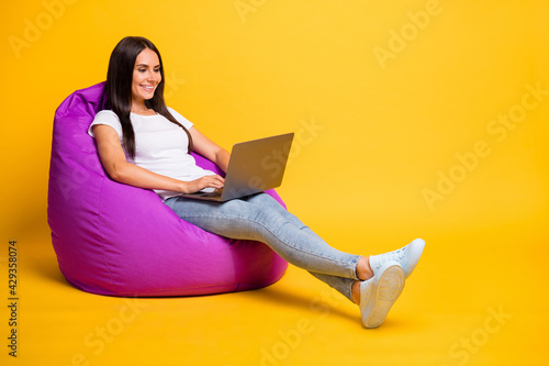 Full length body size photo of pretty brunette working with laptop in purple beanbag isolated on vivid yellow color background