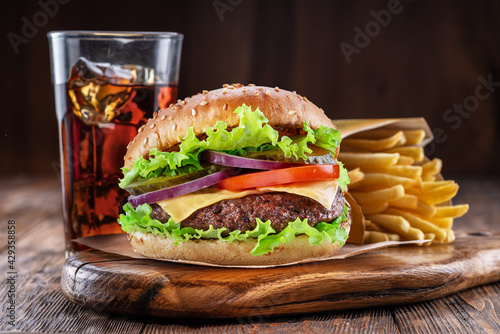 Delicious burger with cola and potato fries on a wooden table with a dark brown background behind. Fast food concept.
