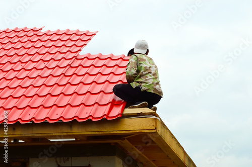 A worker sits on the edge of the roof to attach a sheet of red metal tiles to the boards