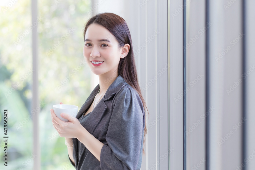 Asian beautiful businesswoman standing by holding a coffee cup by the window happily in the workplace while work from home. Asian beautiful businesswoman standing by holding a coffee cup by the window happily in the workplace while work from home.