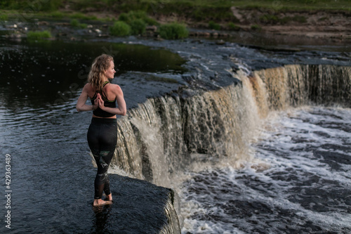 Blonde young woman in sportswear practicing breathing yoga outdoors in harmony with nature. Fitness girl meditating on the edge of waterfall with hands clasped behind her back in namaste mudra sign.