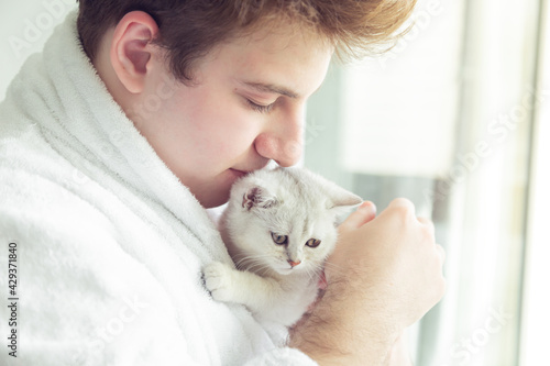 Young caucasian man in a bathrobe is holding a white kitten.
