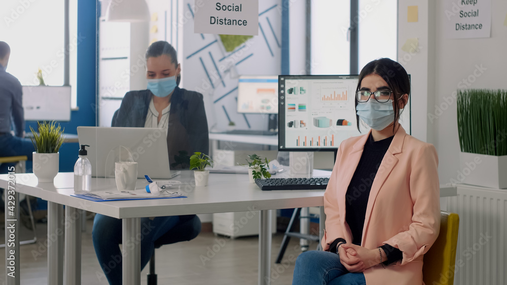 Fototapeta premium Portrait of manager with face mask sitting on chair at desk table in nre normal business company office. Teamworkers working in background respecting social distancing during coronavirus pandemic